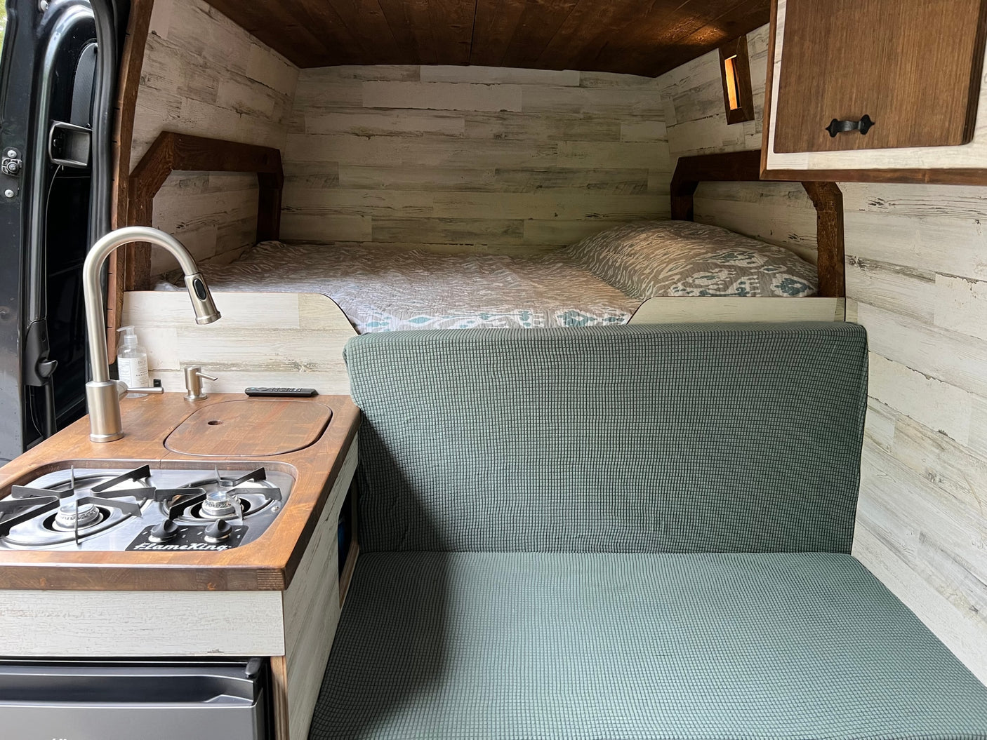 RV rental interior: A cozy camper van featuring a bed, kitchen with stove, and green seating area with wood walls.