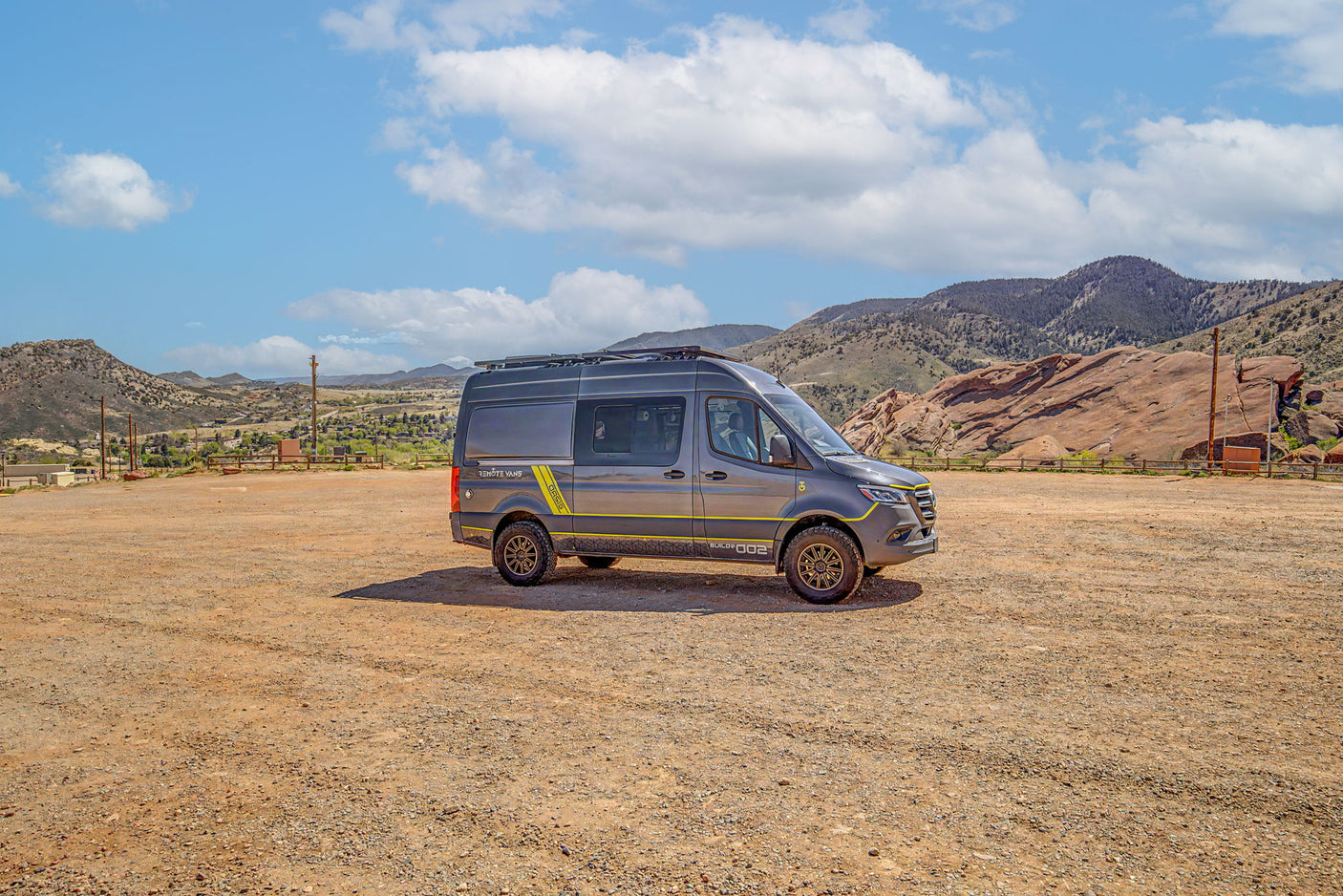 Adventure-ready gray rv rental van with yellow accents in a dirt lot, backed by scenic mountains and blue sky.