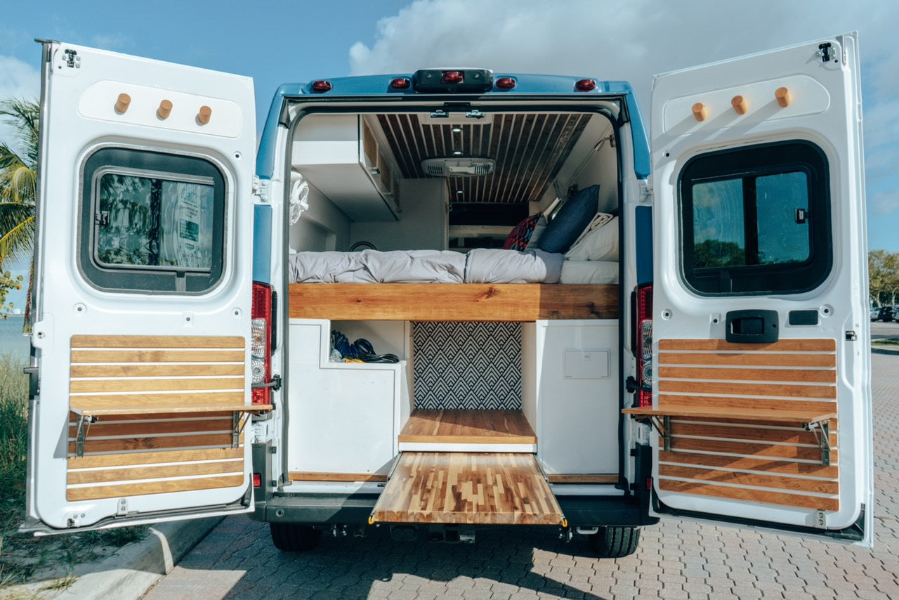 Rear view of an RV rental camper van with open doors, revealing an elevated bed, pull-out table, and custom interior.
