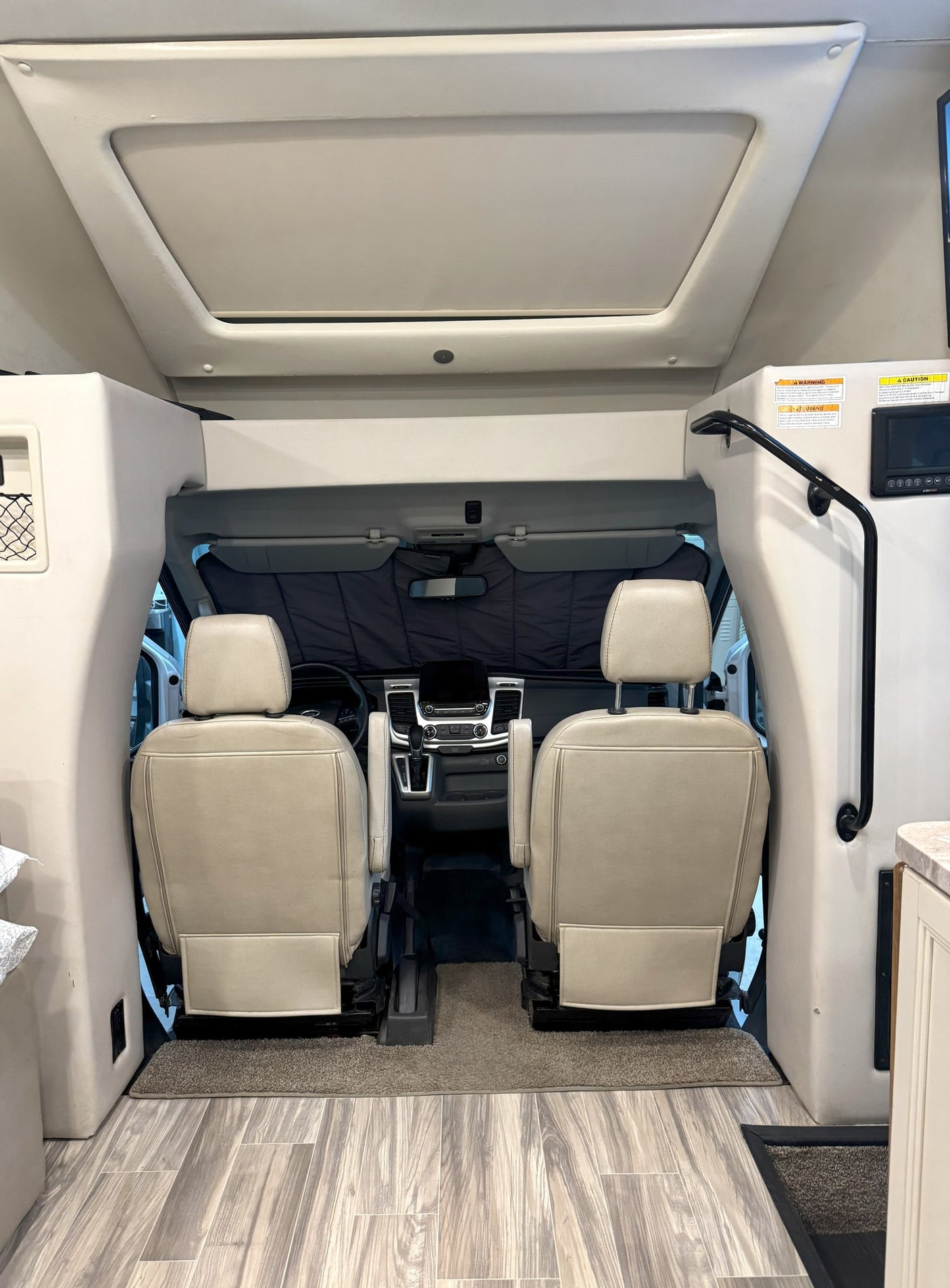 Interior view of an RV rental, showing the cockpit with two tan seats, dashboard, and an overhead bunk/storage area.