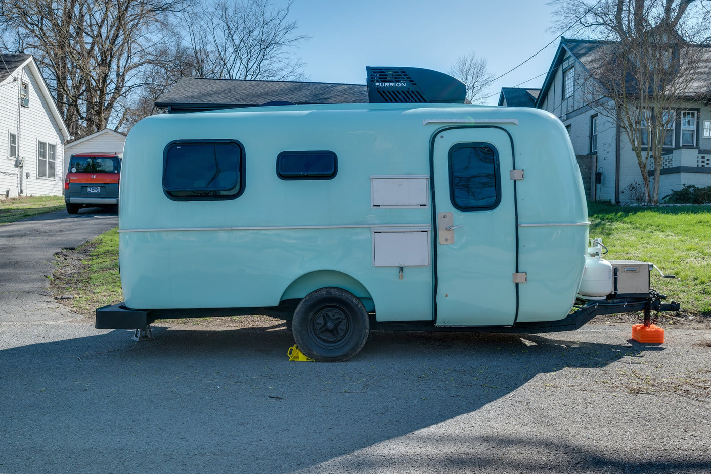 A light blue, rounded teardrop RV rental is parked on asphalt, showing its Furrion AC unit and propane tank.