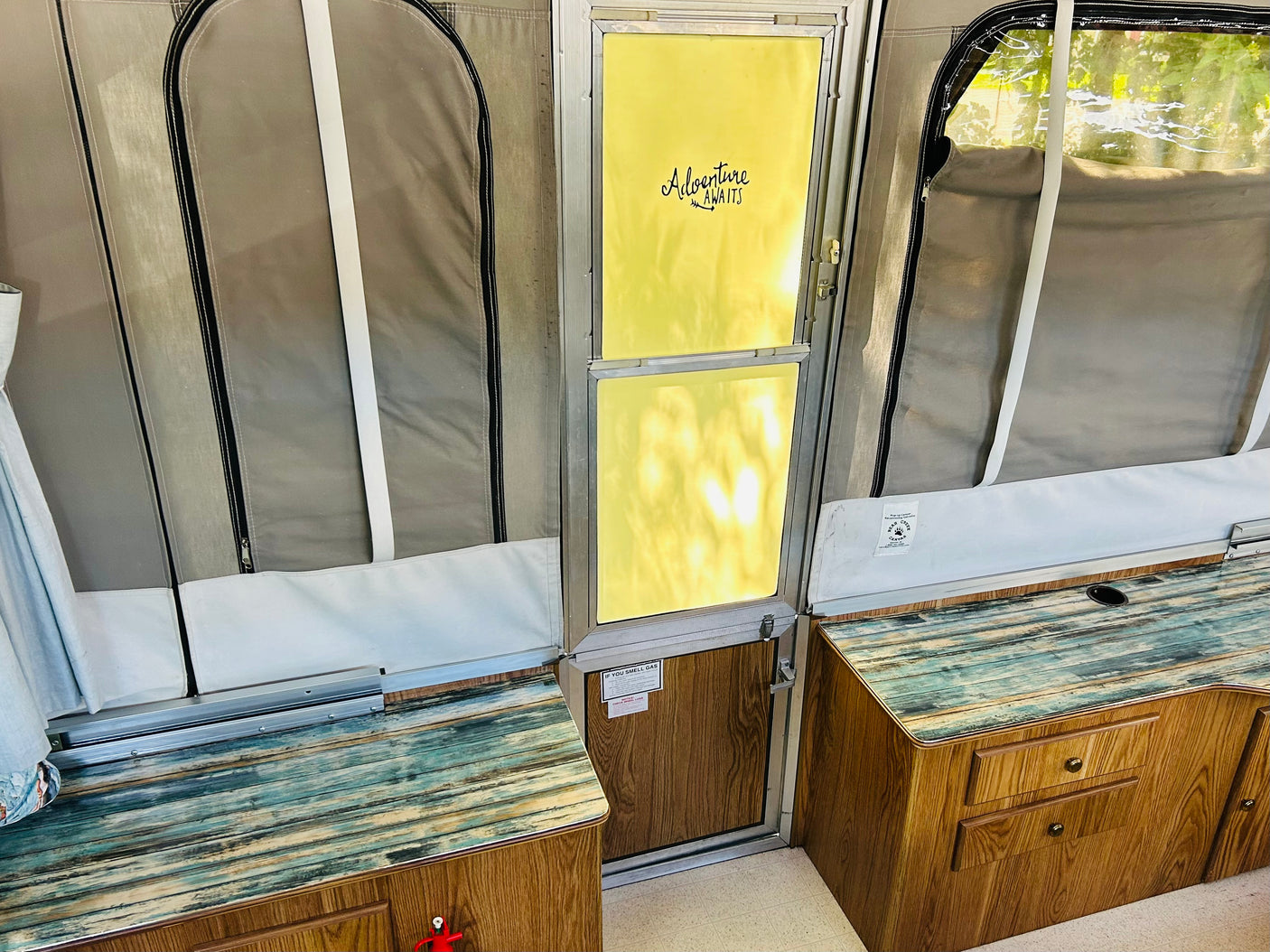 Pop-up camper rv rental interior: fabric walls, wood cabinets, distressed countertops, and "Adventure Awaits" door.