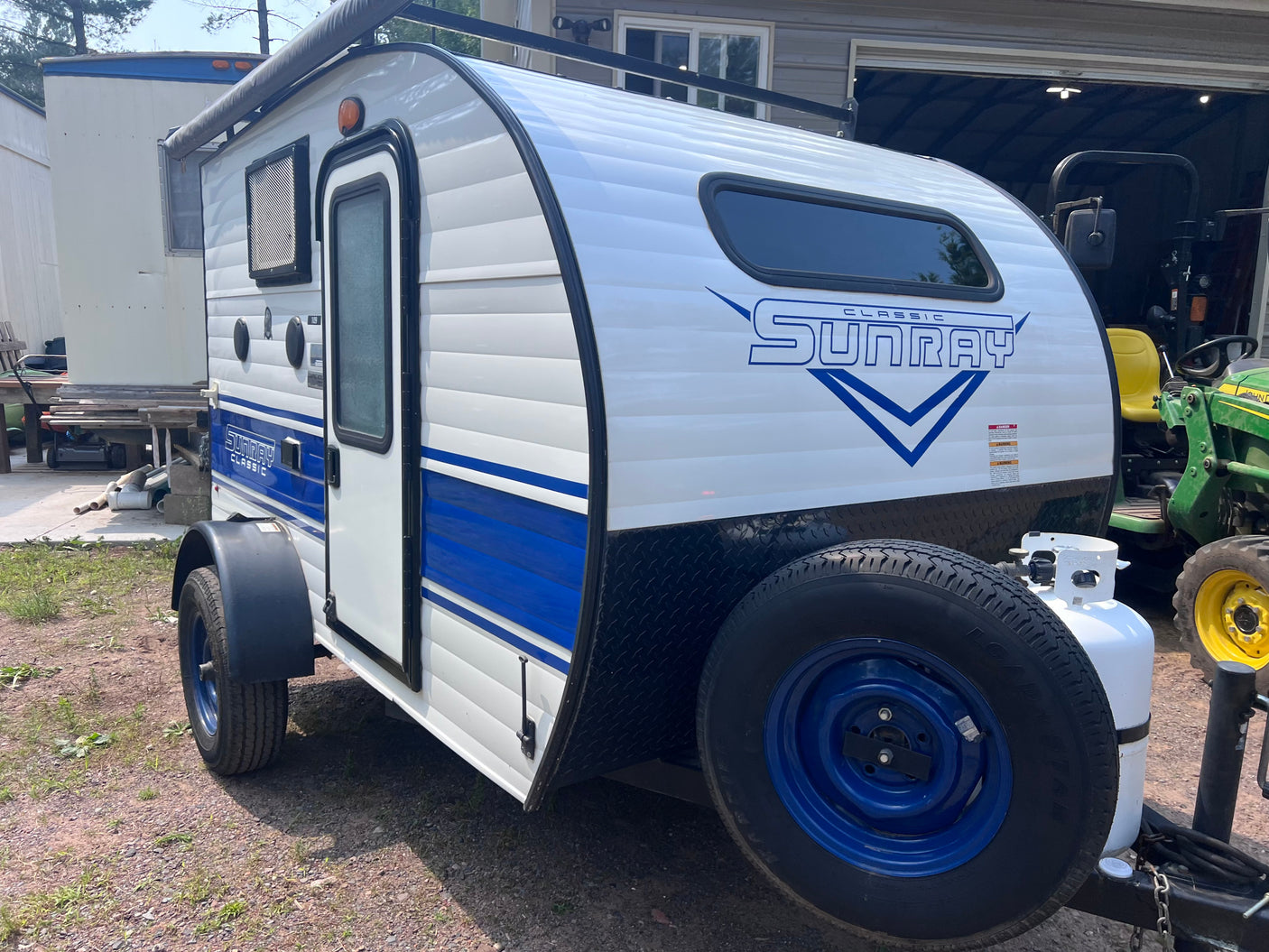 White and blue Classic Sunray teardrop camper, ideal for RV rental, parked outdoors with propane tank & spare tire.