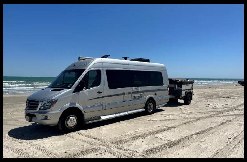 Silver RV rental van & compact trailer on a sunny sandy beach. Ocean waves under clear blue skies, tire tracks visible.