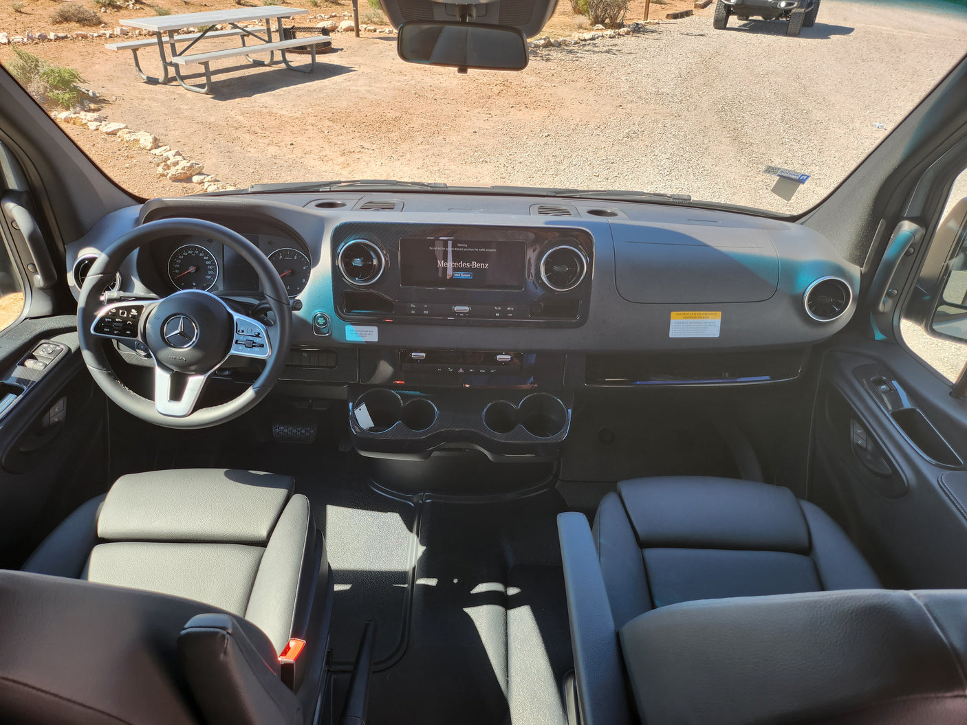 Inside a Mercedes-Benz RV rental: Dashboard, steering wheel, and front seats with a view of a dirt lot and picnic table.