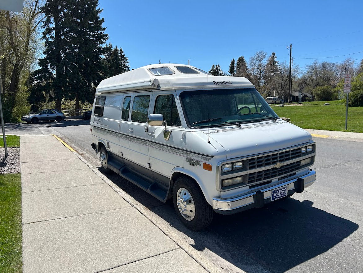 White Roadtrek RV rental (camper van) with high roof, parked on a sunny street next to a sidewalk. Ready for adventure.