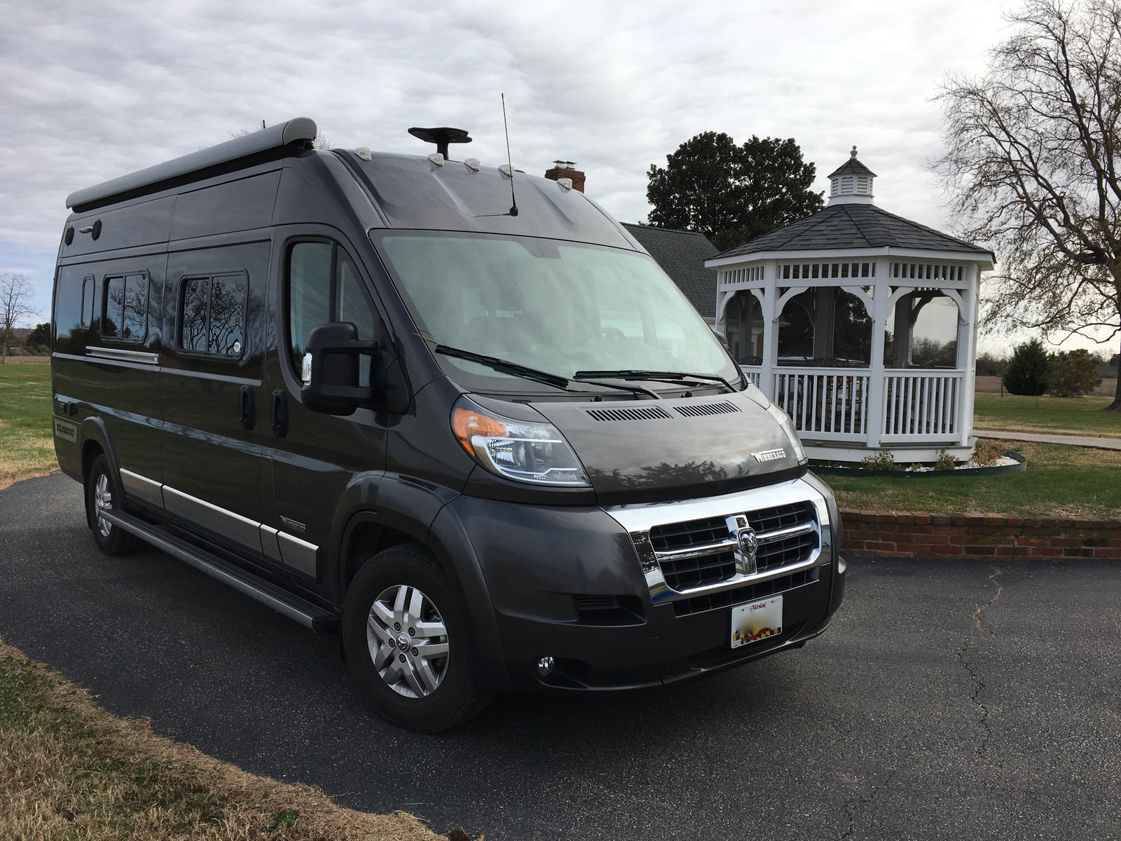 and dark grey exterior.|Sleek dark grey Winnebago Travato RV rental parked on asphalt with a house and bare trees in the background.|A grey Winnebago RV van is parked on asphalt beside a grassy slope leading to a calm bay under a cloudy sky