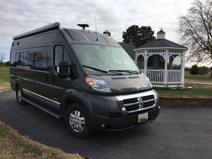 and dark grey exterior.|Sleek dark grey Winnebago Travato RV rental parked on asphalt with a house and bare trees in the background.|A grey Winnebago RV van is parked on asphalt beside a grassy slope leading to a calm bay under a cloudy sky
