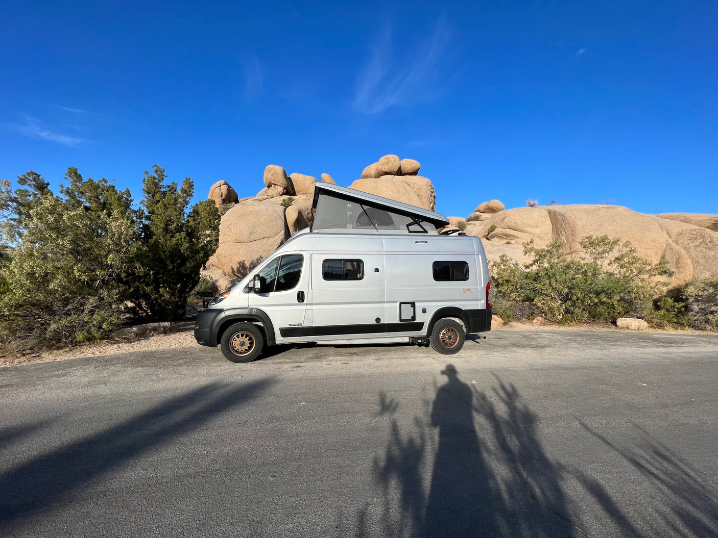 White Winnebago Solis RV rental with pop-top extended, parked among desert rocks and vegetation under a bright blue sky.
