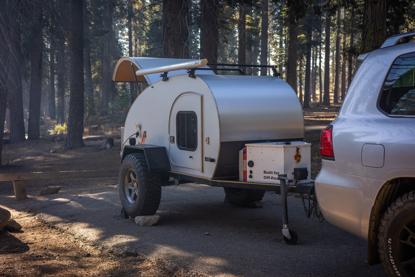 Off-road teardrop camper hitched to SUV in a sunny forest setting, ideal for adventure travel or an RV rental.