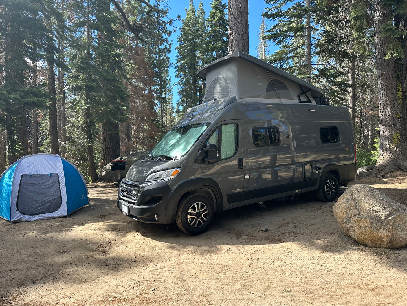 RV rental camper van with pop-top roof and blue tent at a sunny forest campsite. A large rock is nearby on the dirt ground.