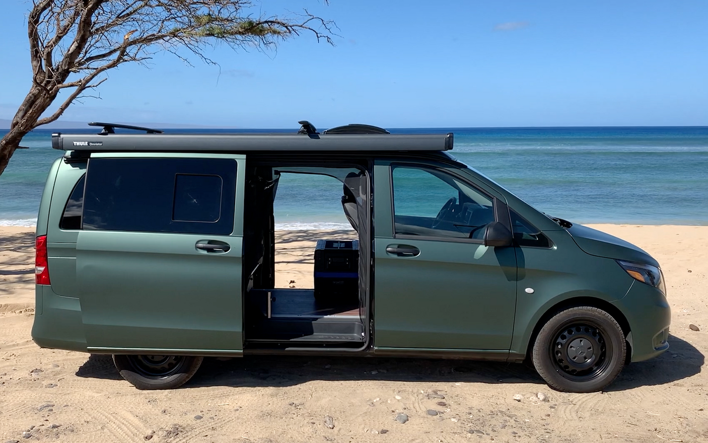 Green camper van, equipped with a Thule awning, parked on a sandy beach overlooking the ocean. Ideal for RV rental.