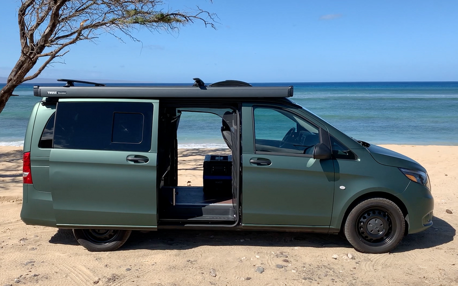 Green camper van, equipped with a Thule awning, parked on a sandy beach overlooking the ocean. Ideal for RV rental.
