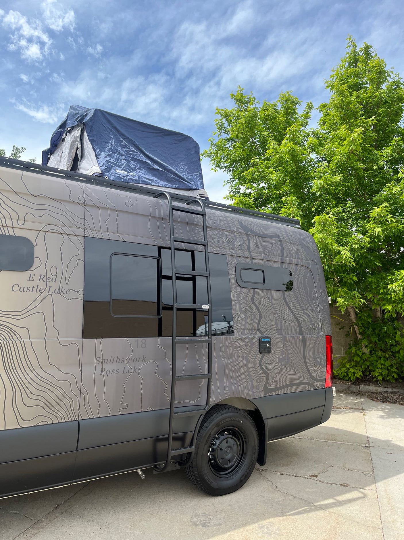 This custom rv rental camper van, adorned with a topographical map design, features a ladder and rooftop tent, ideal for adventure travel.