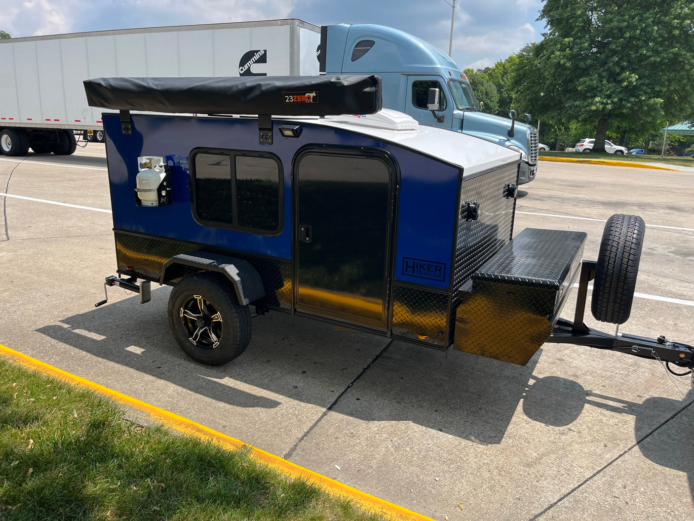 A blue Hiker Teardrop Trailer, a compact RV rental, with an awning and propane tank, parked on asphalt.