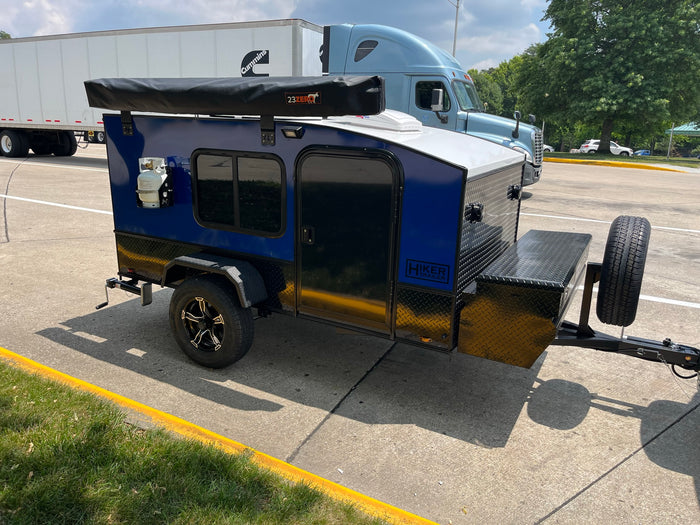 A blue Hiker Teardrop Trailer, a compact RV rental, with an awning and propane tank, parked on asphalt.