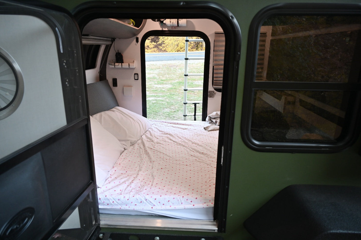 Inside an RV rental: a compact sleeping area with a polka dot comforter. The open rear hatch reveals a grassy outdoor view.