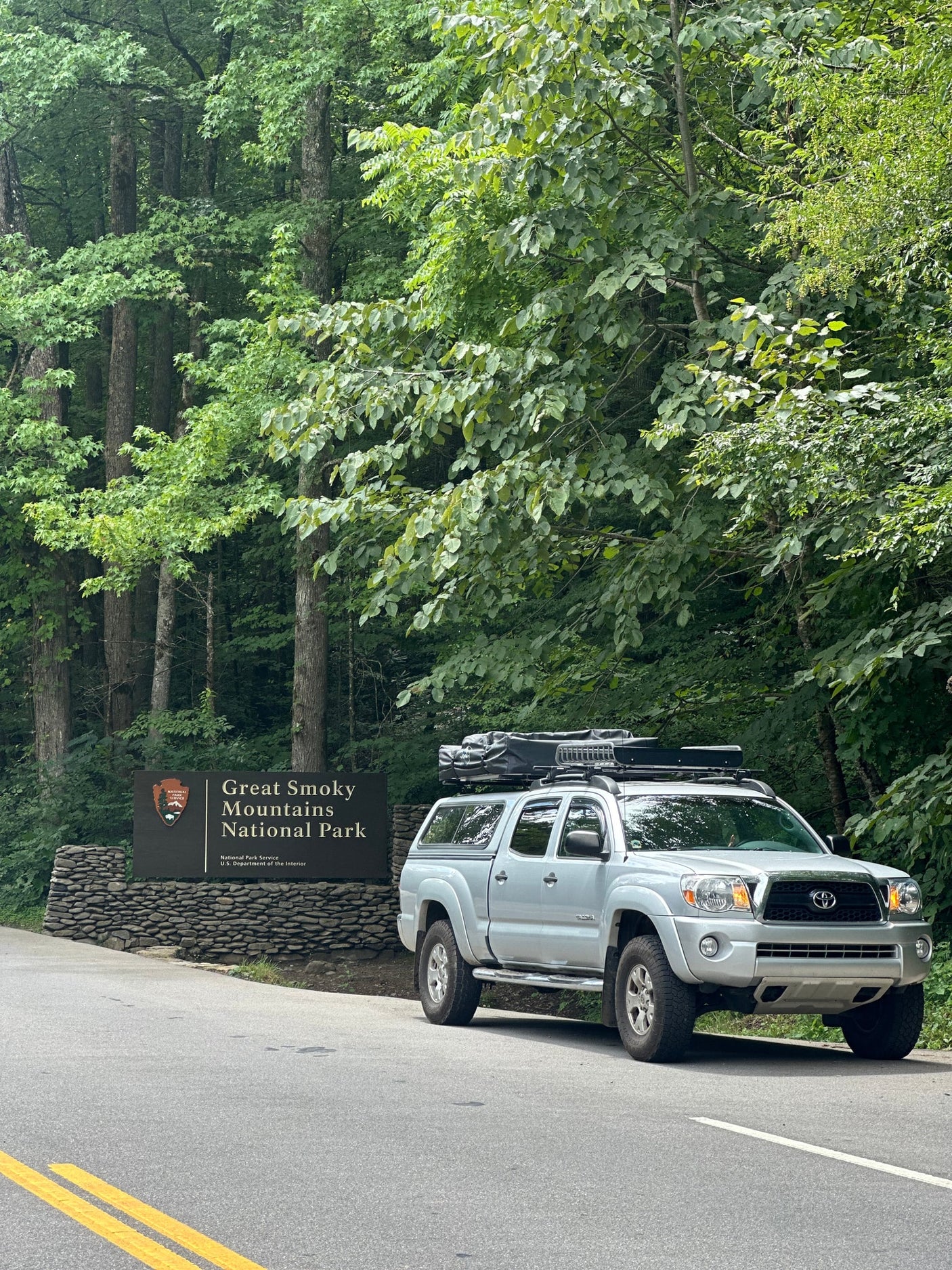 Enjoy nature! This truck with a rooftop tent offers a unique alternative to an RV rental for camping in the woods.|A silver pickup truck with a TMBK rooftop tent set up for camping in a lush forest campsite