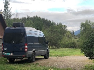 Experience adventure! This dark camper van RV rental is parked in a picturesque mountain landscape with forest & cabin nearby.