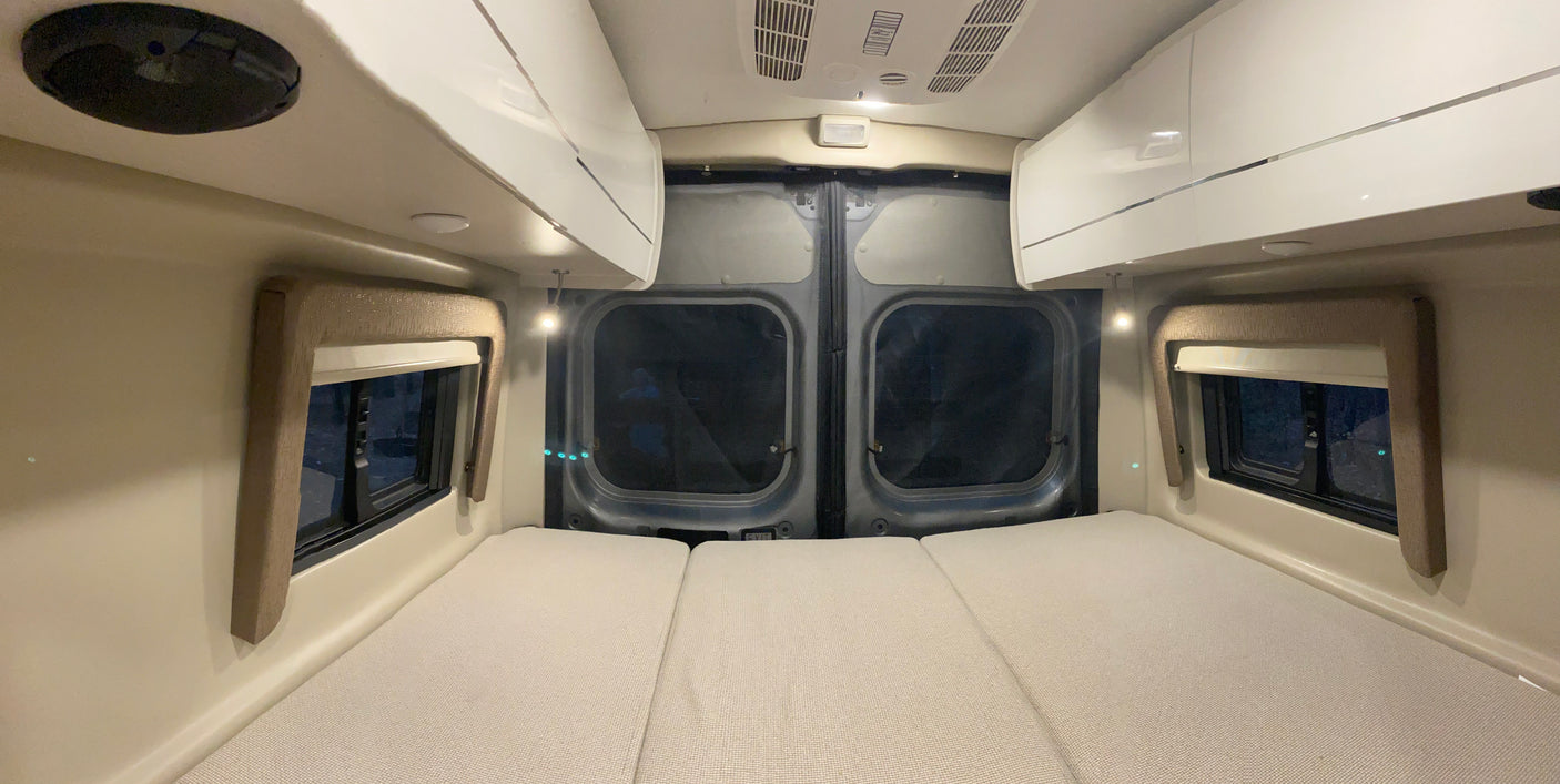 Inside an RV rental: a cozy sleeping area with three laid-out mattresses, side windows, and overhead cabinets.