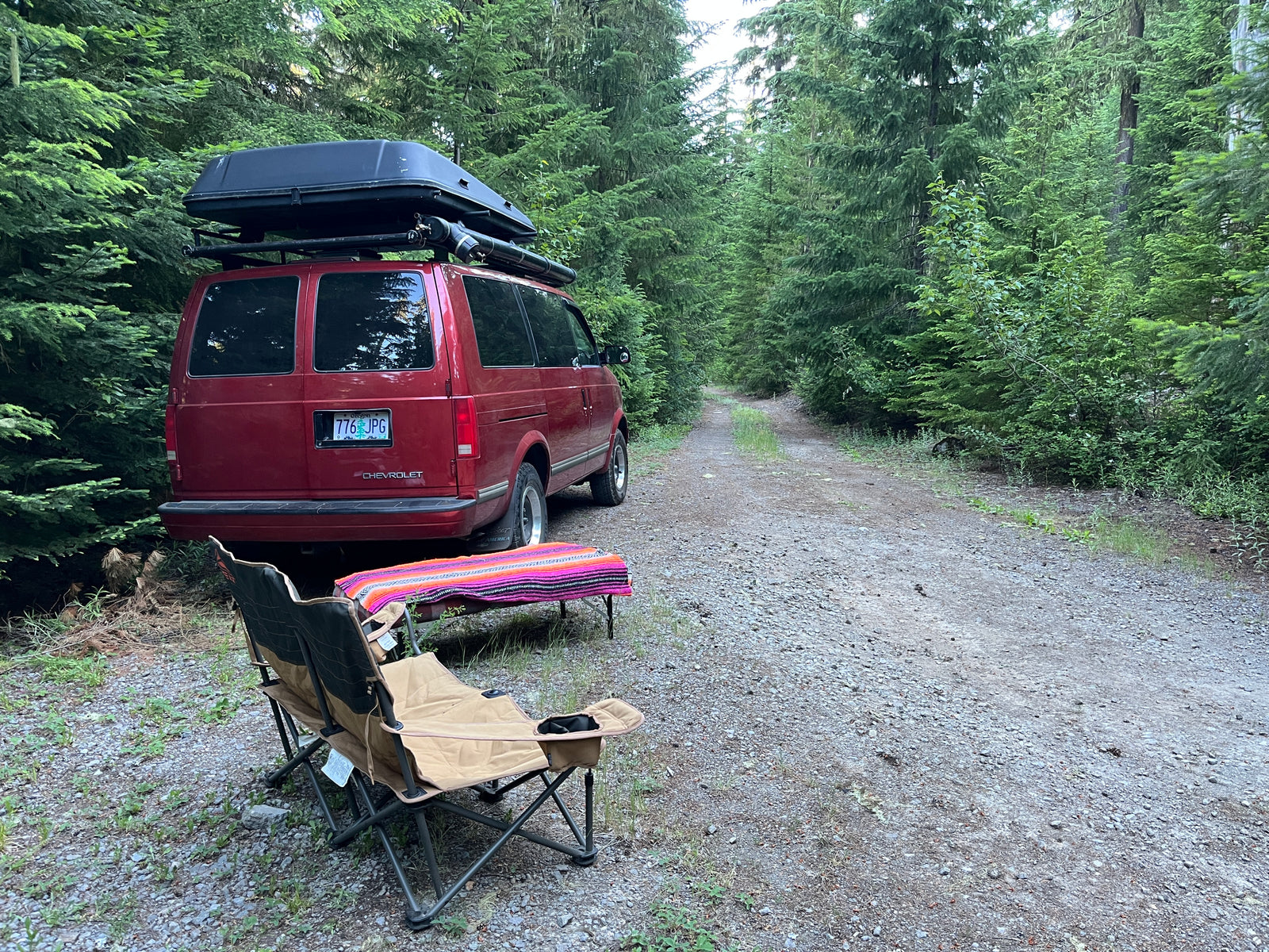 Red camper van with roof box on a forest road, chairs & table set up for off-grid RV rental camping.