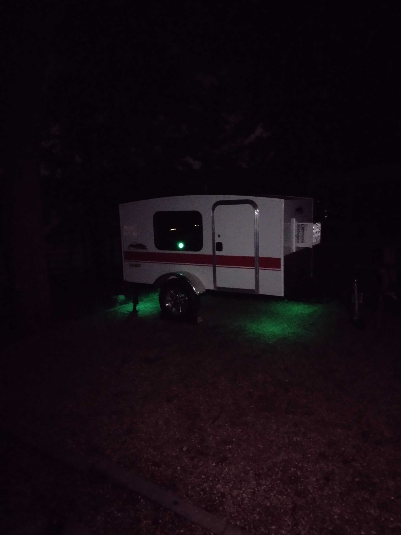 Night view of a compact white and red rv rental trailer, illuminated by vibrant green underglow lights, parked outdoors.