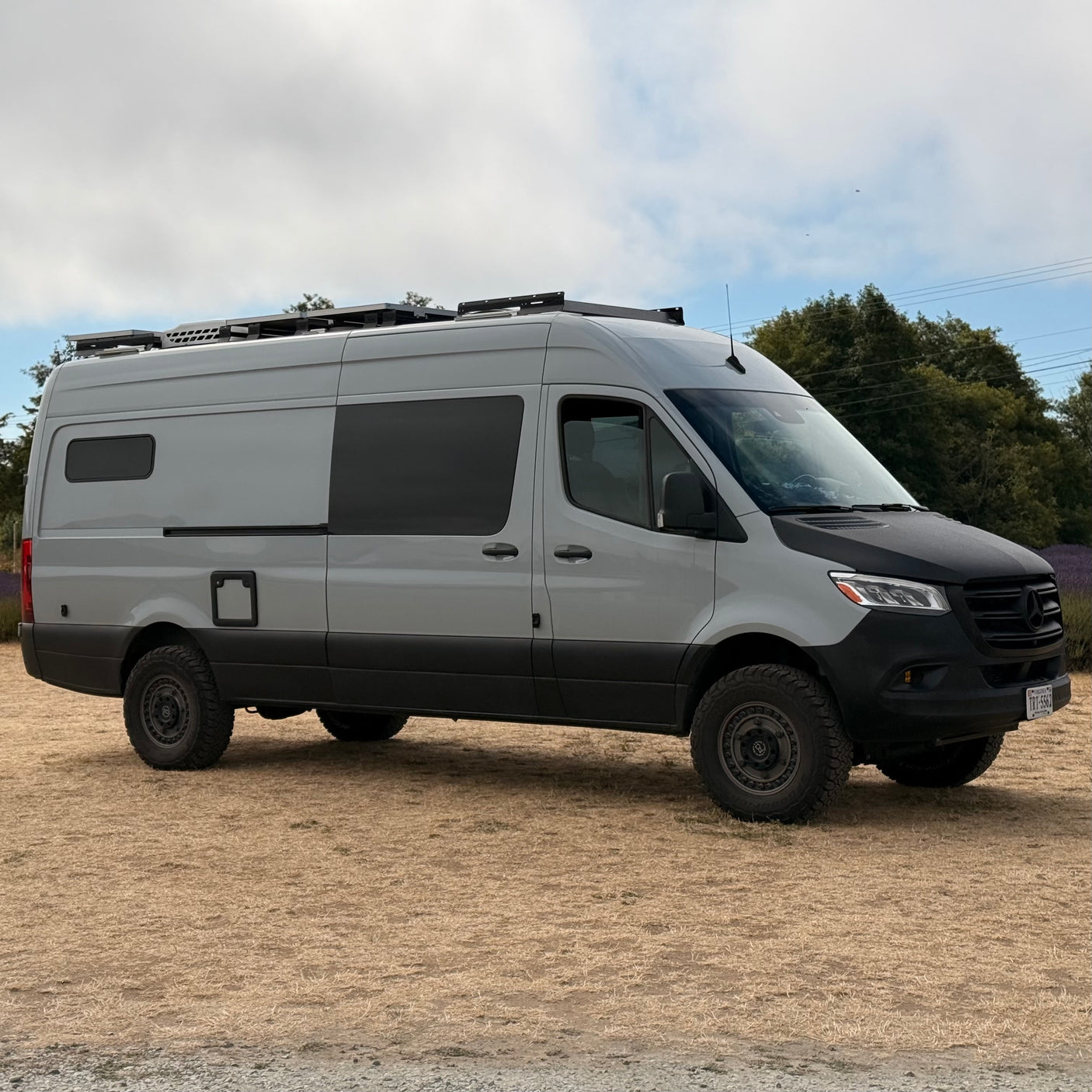 Rugged grey Mercedes Sprinter camper van with roof rack, ideal for RV rental adventures, parked on dry ground.
