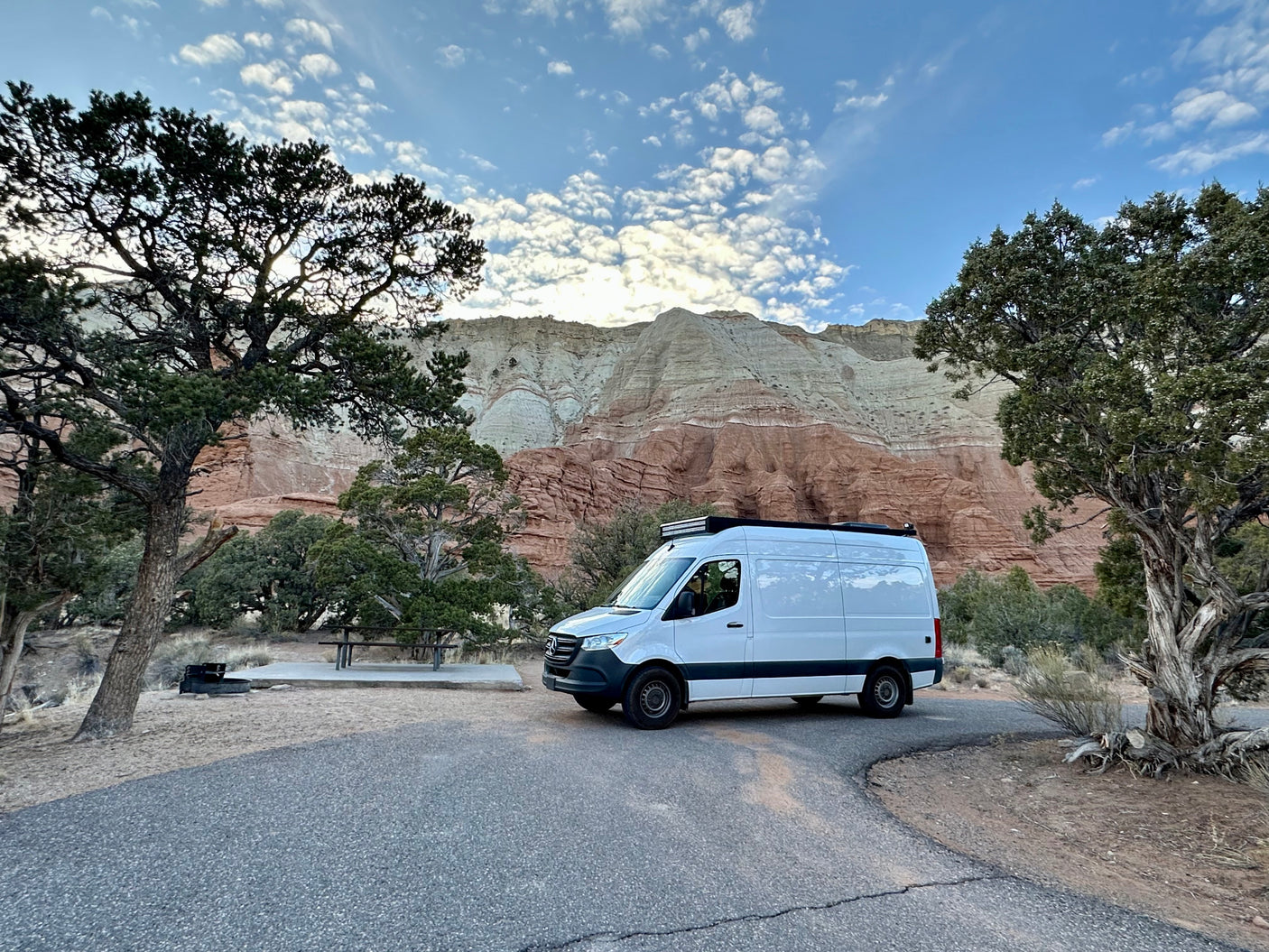 White RV rental van at a scenic campsite, framed by layered mountains under a cloudy sky.