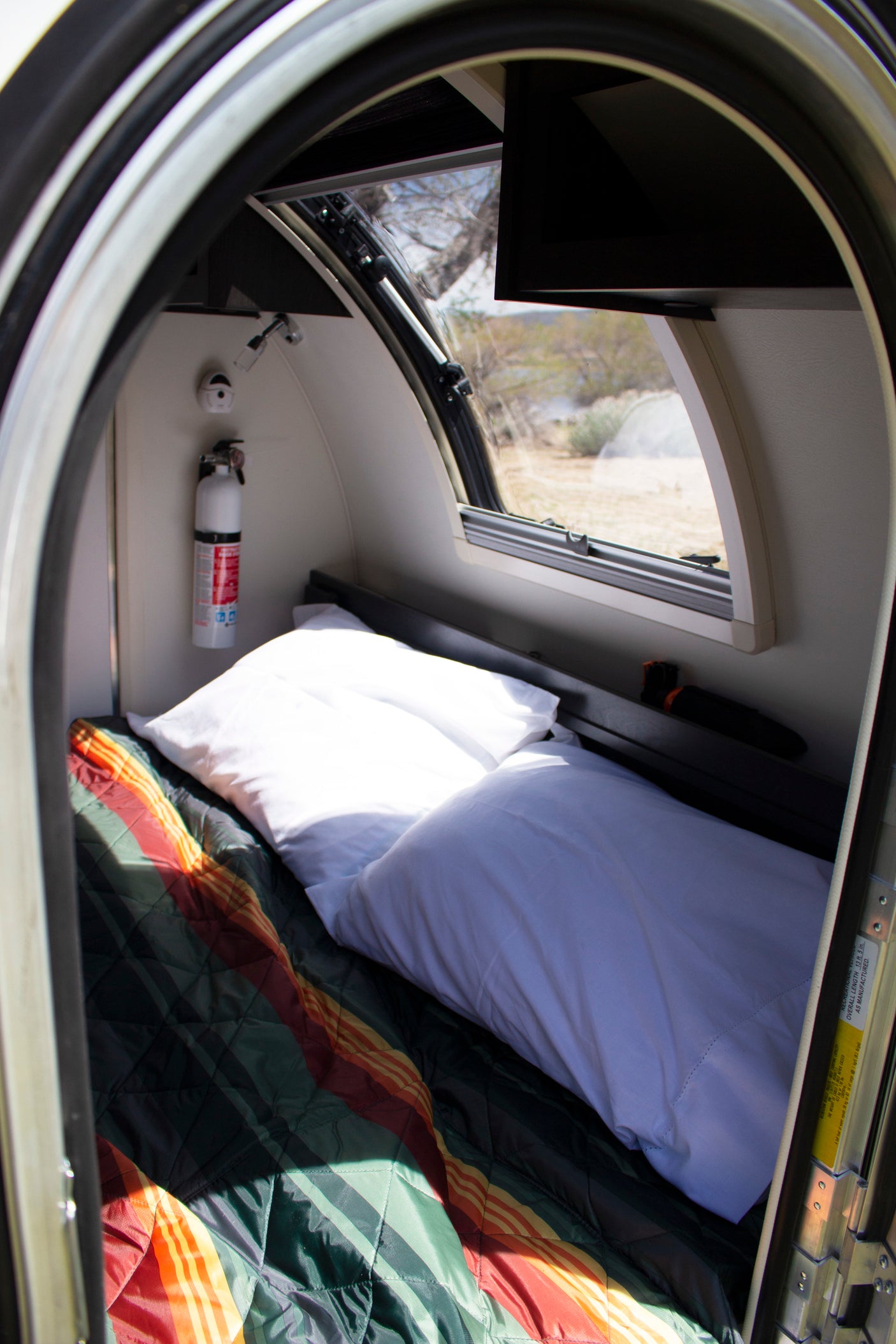 Inside a compact RV rental: a cozy bed, fire extinguisher, and window with scenic desert views.