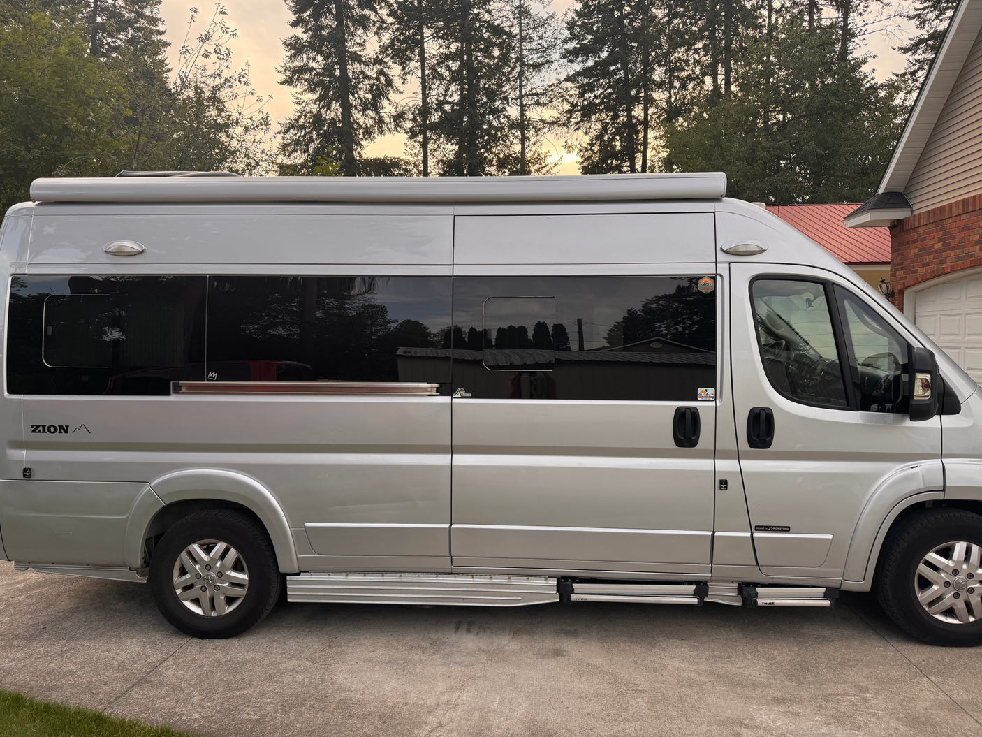 Side view of a silver Roadtrek Zion camper van, perfect for an RV rental. It's parked on concrete with trees and a house in the background.