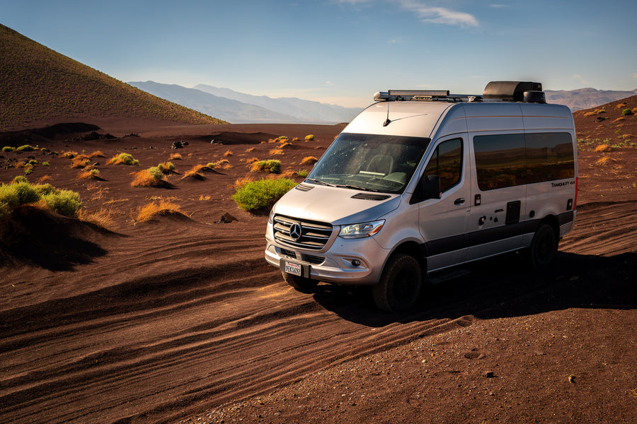 Silver Mercedes Sprinter **rv rental** van on dark volcanic sand with desert plants and distant mountains under a clear sky.