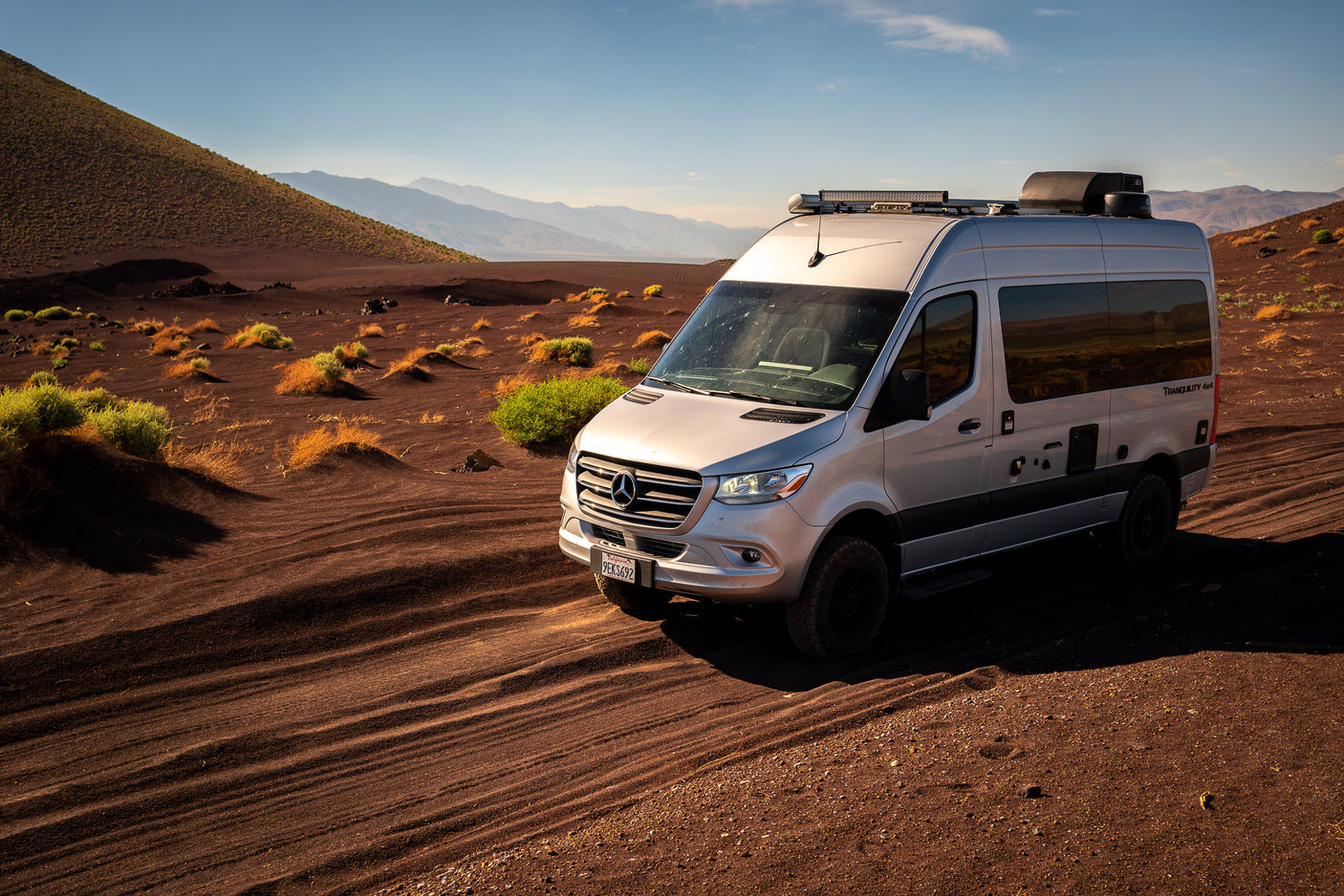 Silver Mercedes Sprinter **rv rental** van on dark volcanic sand with desert plants and distant mountains under a clear sky.