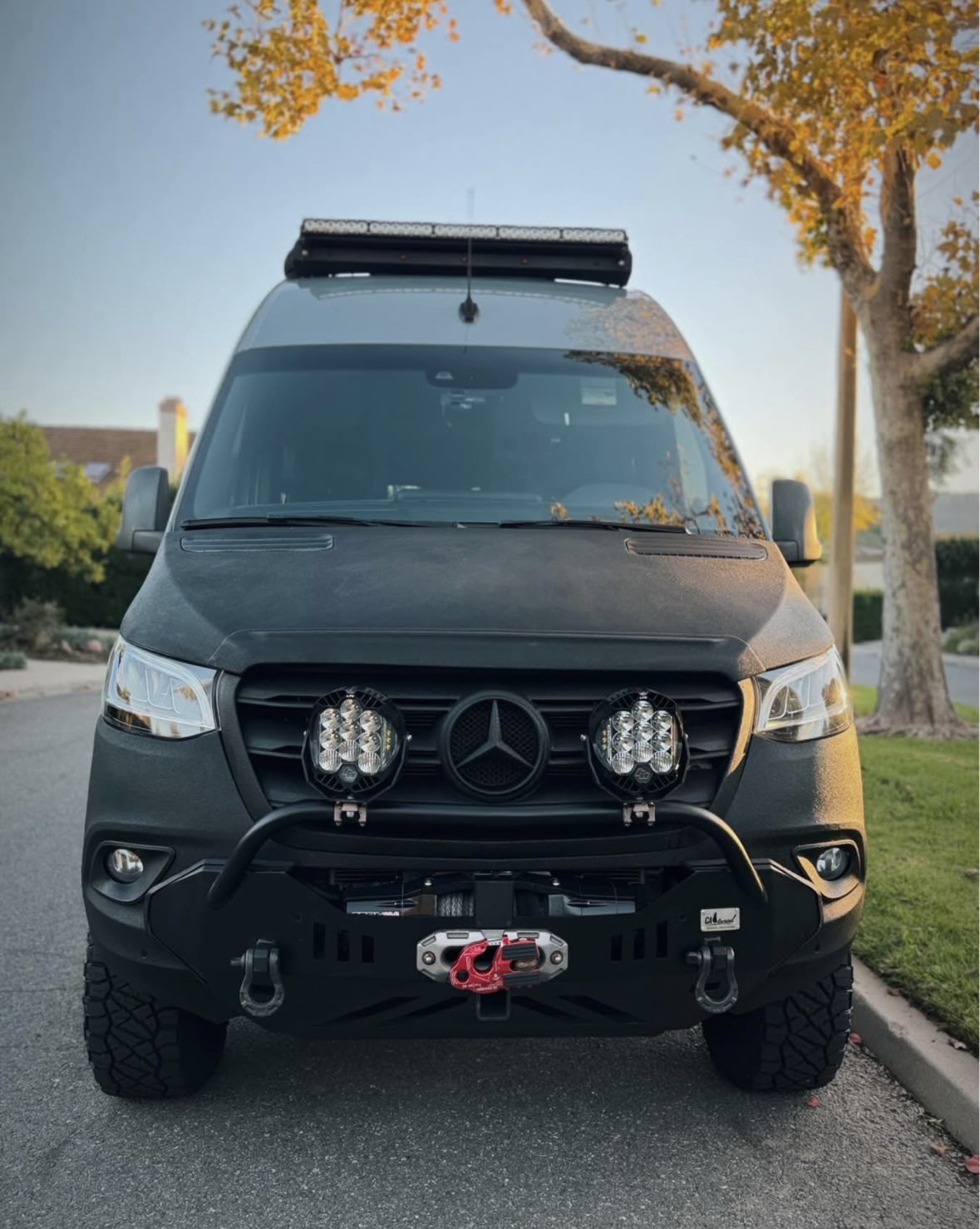 This custom Mercedes Sprinter off-road RV rental boasts a robust front bumper, winch, and roof light bar, parked under autumn leaves. — photo 7