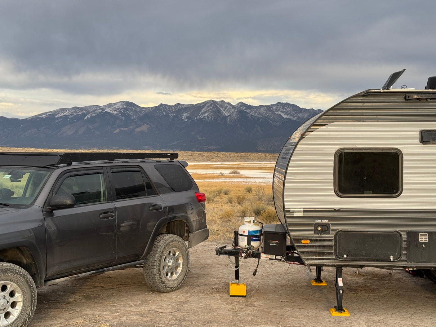 An RV rental & dark SUV parked in a vast, dry landscape with snow-dusted mountains under a dramatic sky, ready for outdoor adventure.