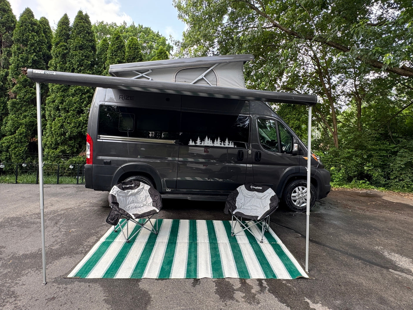 Stylish dark grey Rize camper van with pop-top roof and Thule awning. An outdoor mat & chairs complete this RV rental setup.