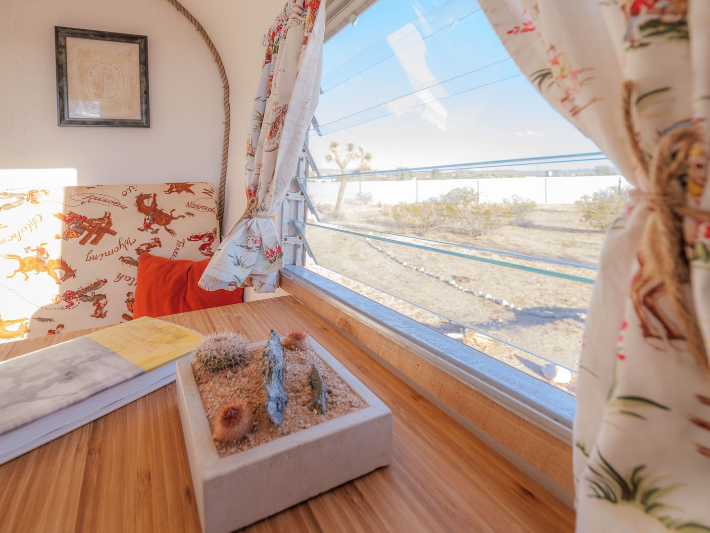 Inside a cozy RV rental: wooden table, desert garden planter, and a window with a clear view of the arid landscape & Joshua Tree.