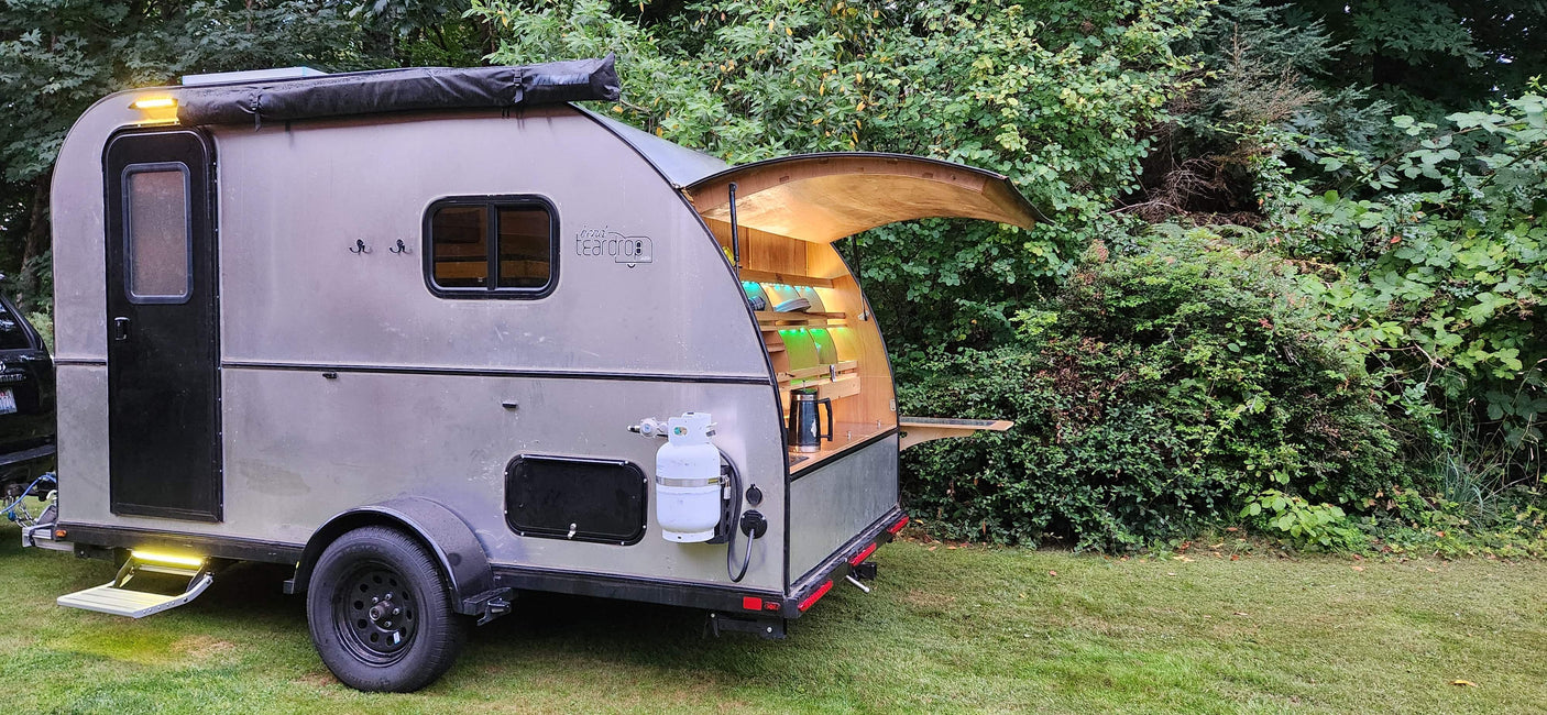 Teardrop RV rental parked in lush greenery, showing its open rear galley kitchen and side features.