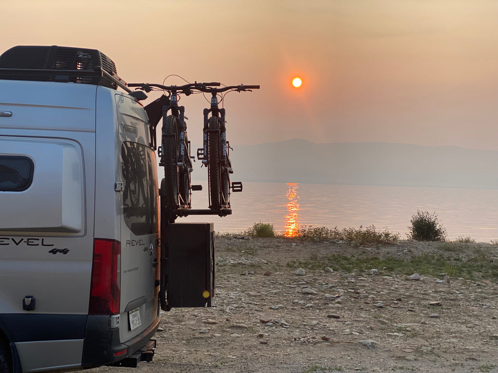 Revel 4x4 RV rental with bikes parked lakeside at a hazy sunset. Golden sun reflects on calm water, hills in distance.