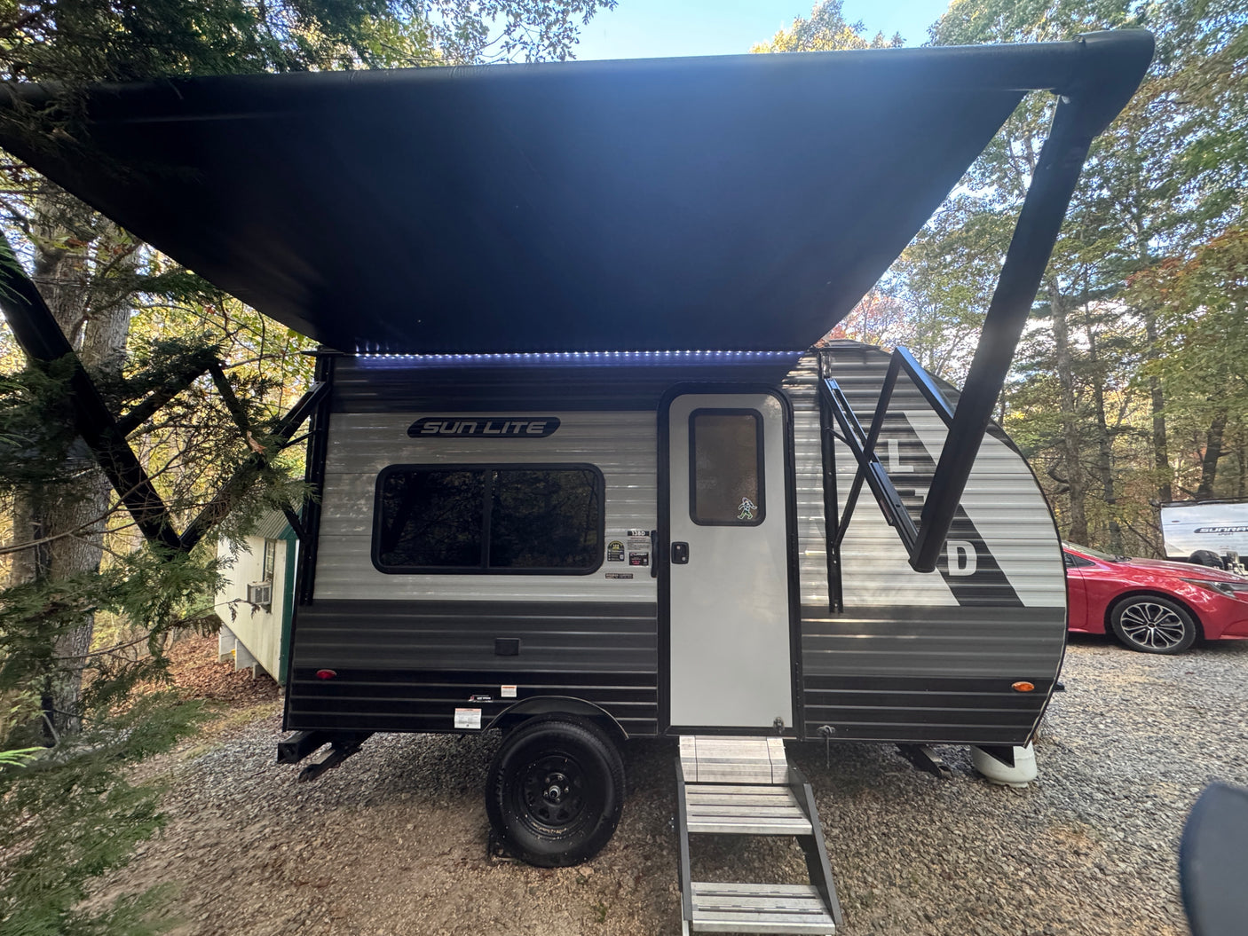 Enjoy this RV rental! A Sun Lite travel trailer with an illuminated awning is set up on gravel in a wooded camp spot.