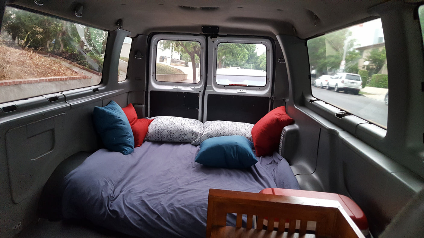 Cozy RV rental interior with a full bed & pillows. Views out side & back windows of parked cars & trees.