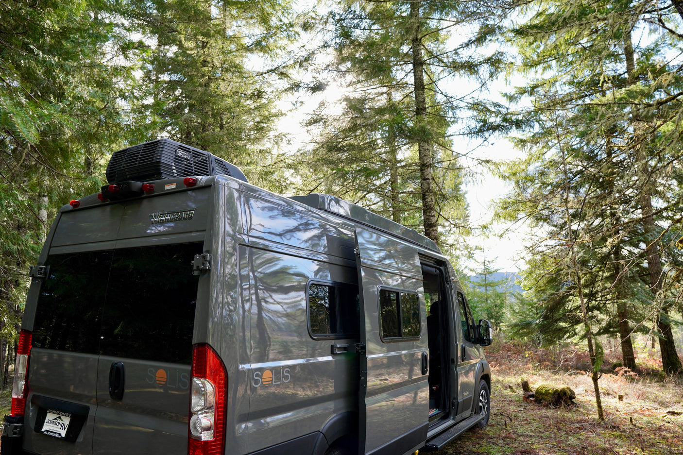 Grey Winnebago Solis RV rental parked in a sun-dappled forest. Side door open for outdoor adventure.