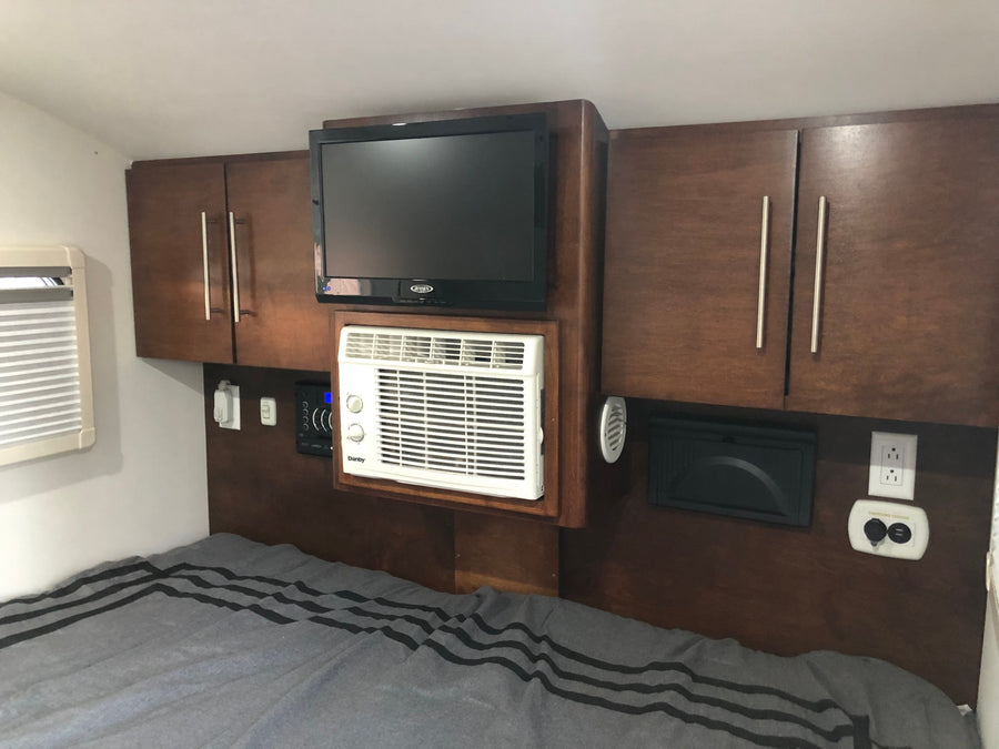 Cozy RV rental bedroom featuring a wall-mounted TV, compact AC unit, cabinets, and a charging center above a striped bed.