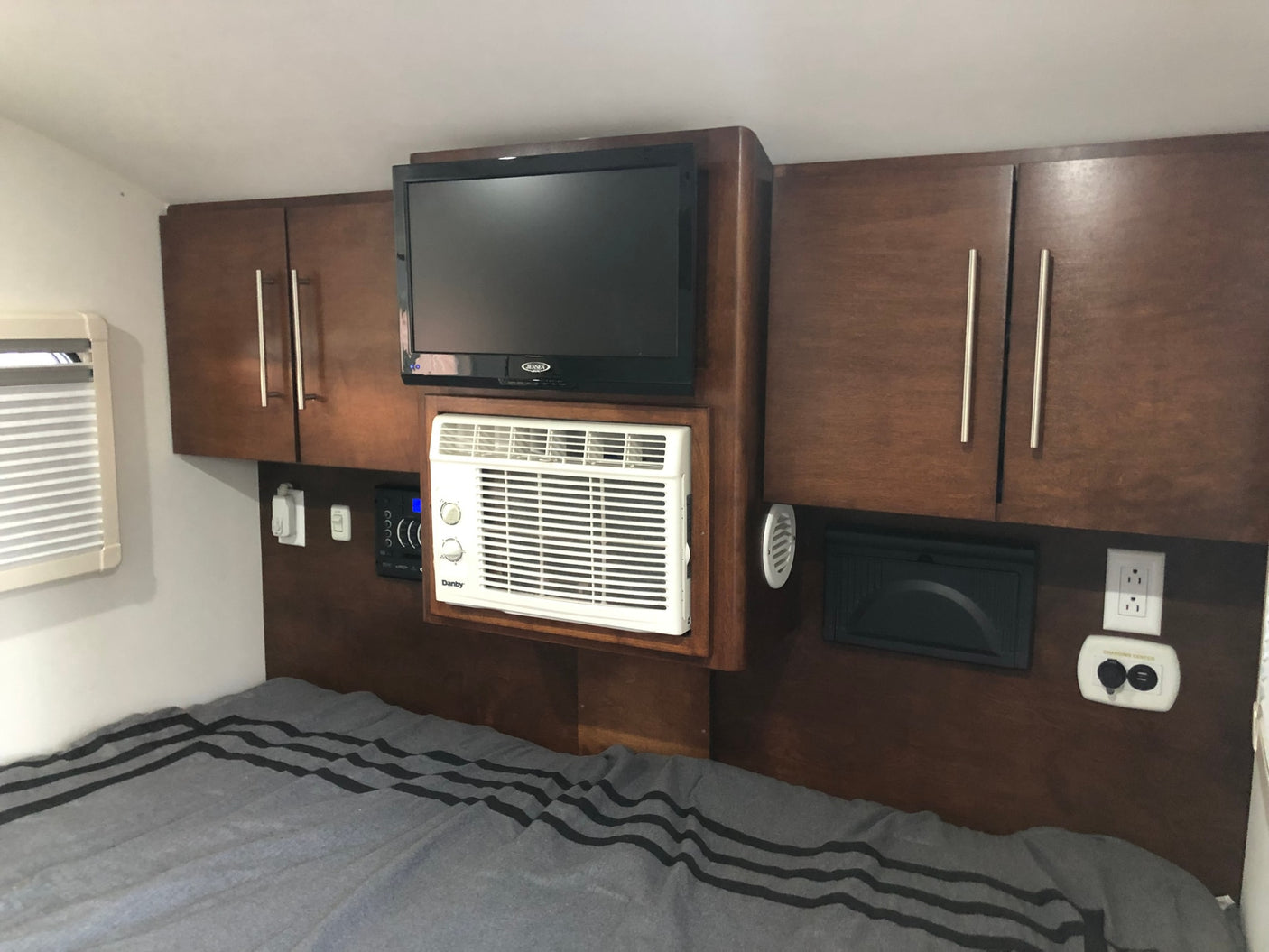 Cozy RV rental bedroom featuring a wall-mounted TV, compact AC unit, cabinets, and a charging center above a striped bed.