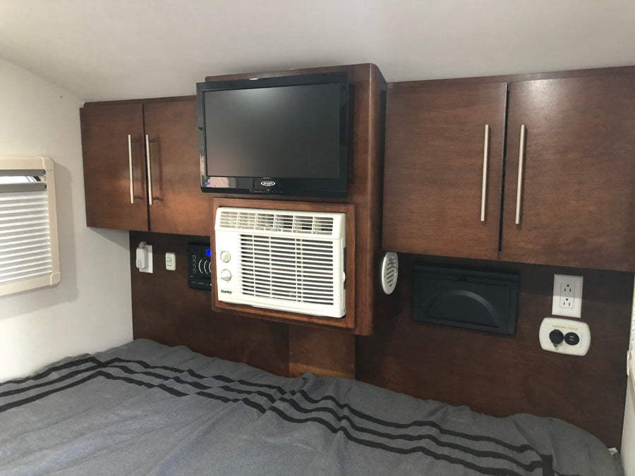 Cozy RV rental bedroom featuring a wall-mounted TV, compact AC unit, cabinets, and a charging center above a striped bed.