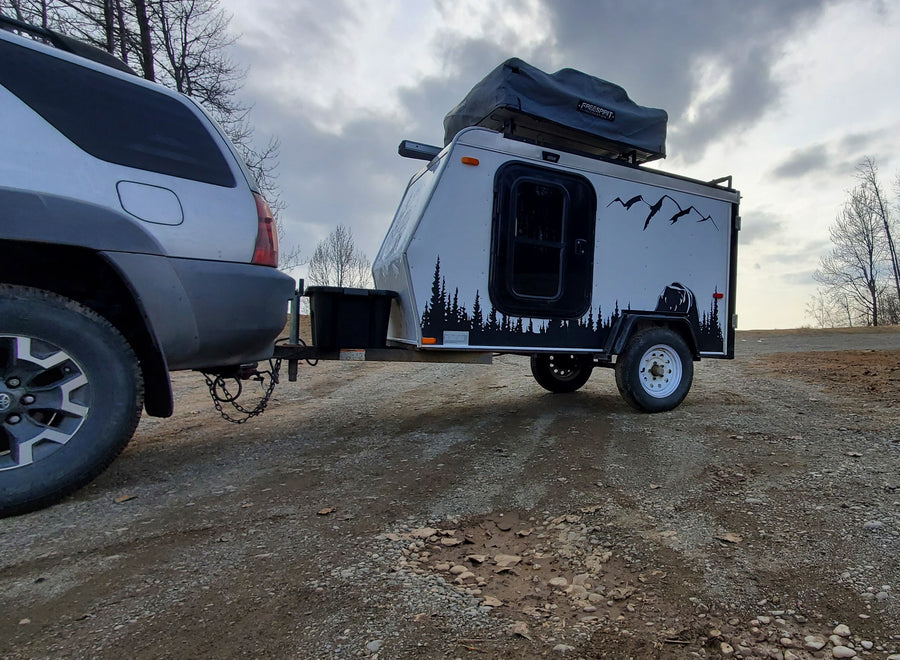 An SUV tows an RV rental teardrop camper with a rooftop tent, ready for outdoor travel on a dirt road.