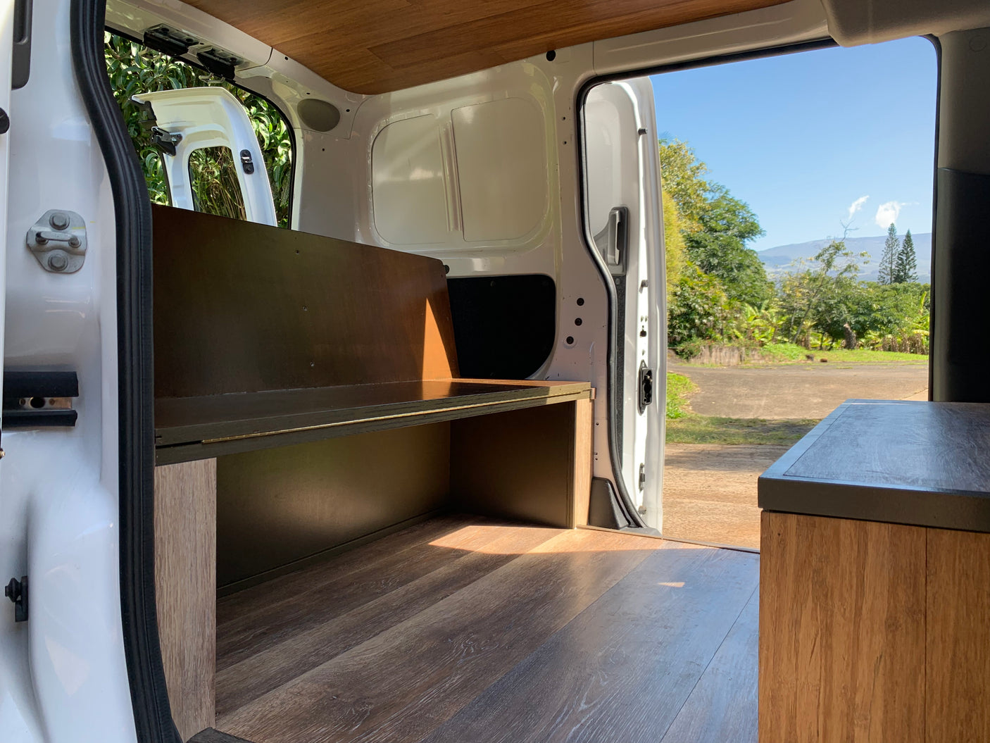 RV rental: Bright interior of a custom camper van with wood floors/ceiling and built-in benches, opening to a lush outdoor scene.
