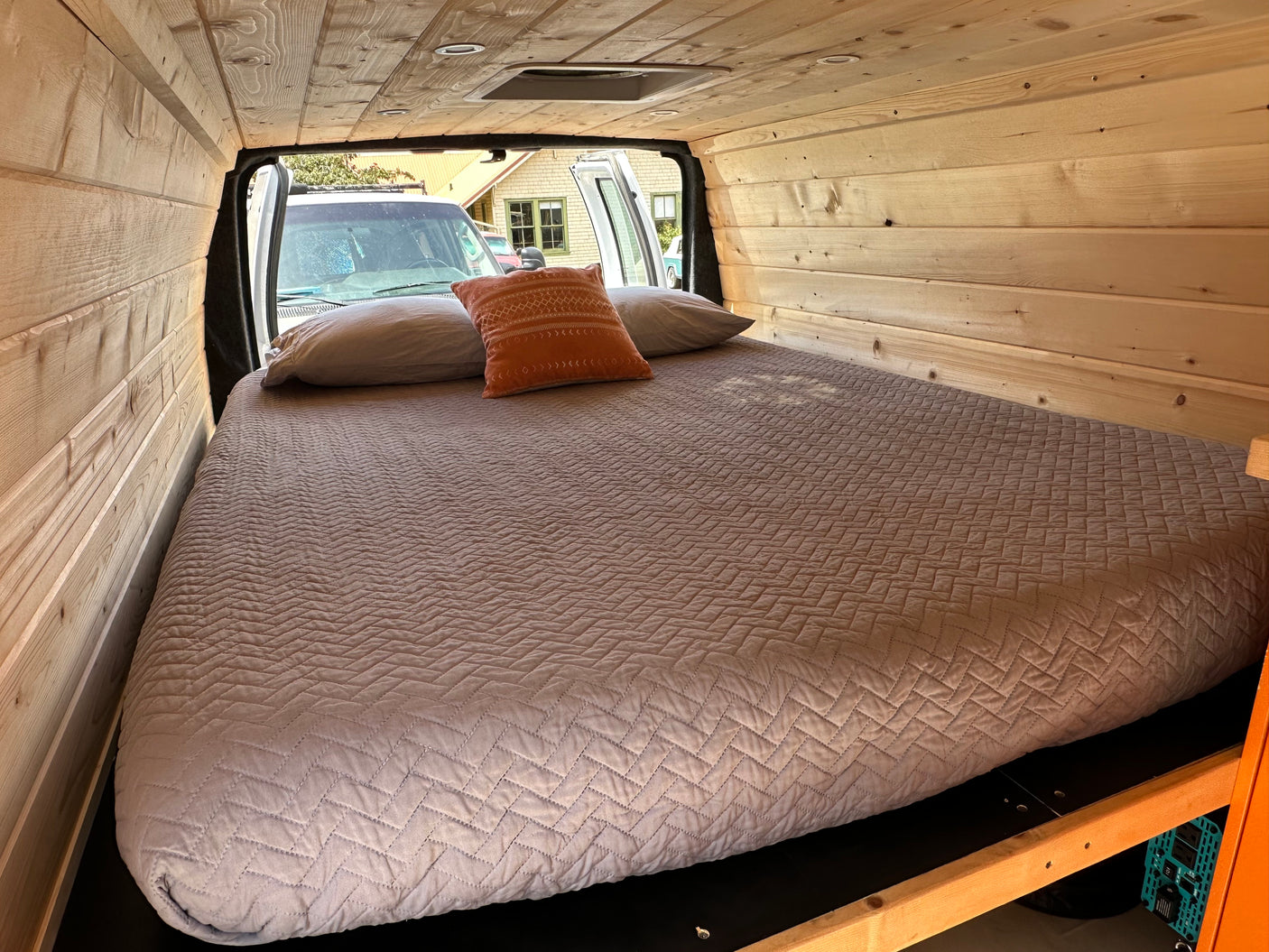 Custom wood paneled RV rental interior with a large comfortable bed and pillows. Rear doors are open, showing the exterior.