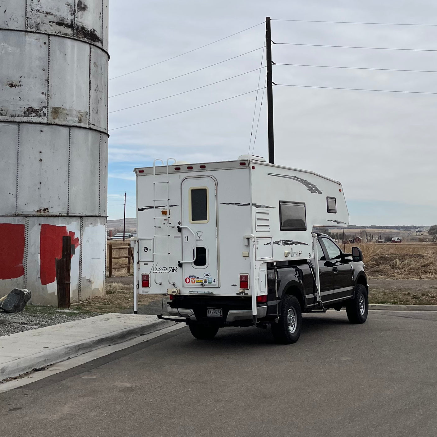 White Northstar truck camper on a black pickup, an ideal RV rental, parked by a metal silo in a rural landscape.