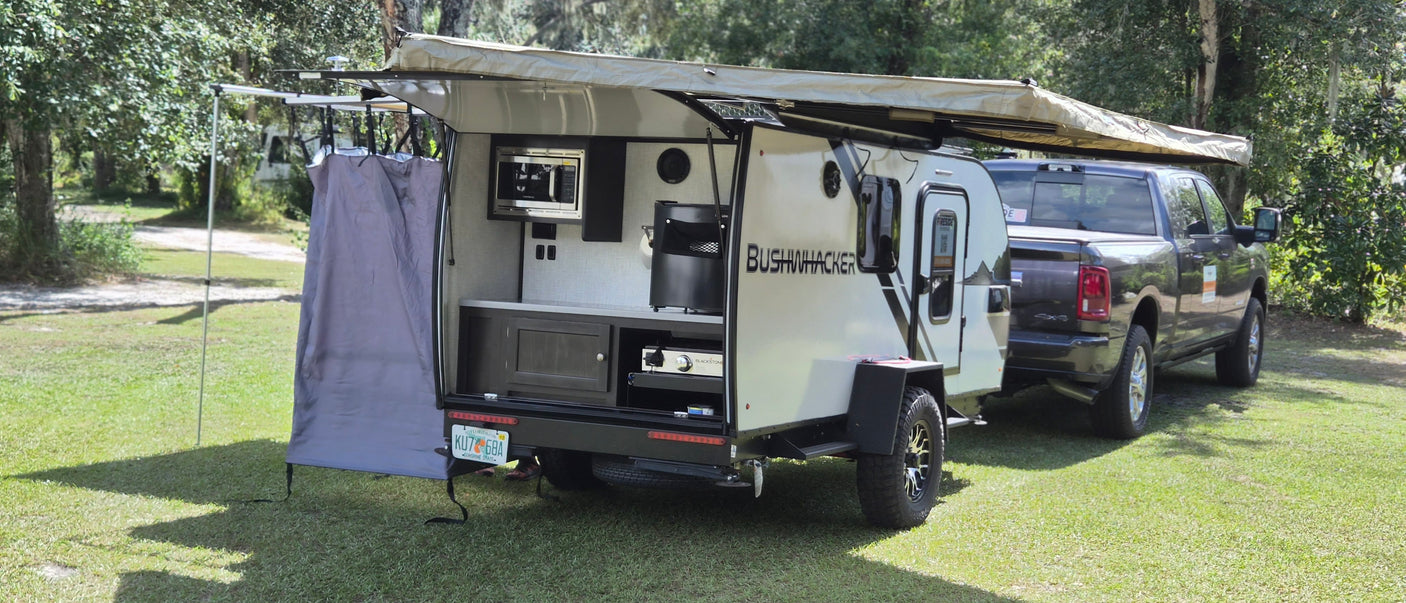 This Bushwhacker travel trailer RV rental with an open outdoor kitchen and awning, hitched to a truck, is set for camping.