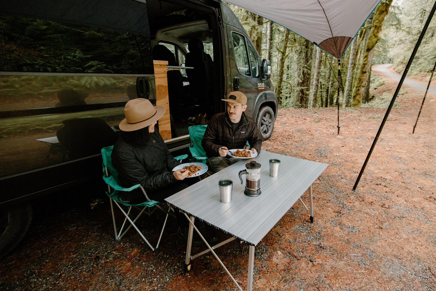 Two people enjoying an outdoor meal from their RV rental under an awning in a lush forest. Perfect for van life.